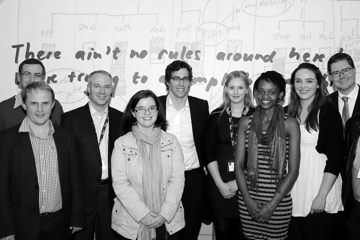 About twenty guests stand together in front of a wall covered in handwritten notes that read "There ain't no rules around here"
